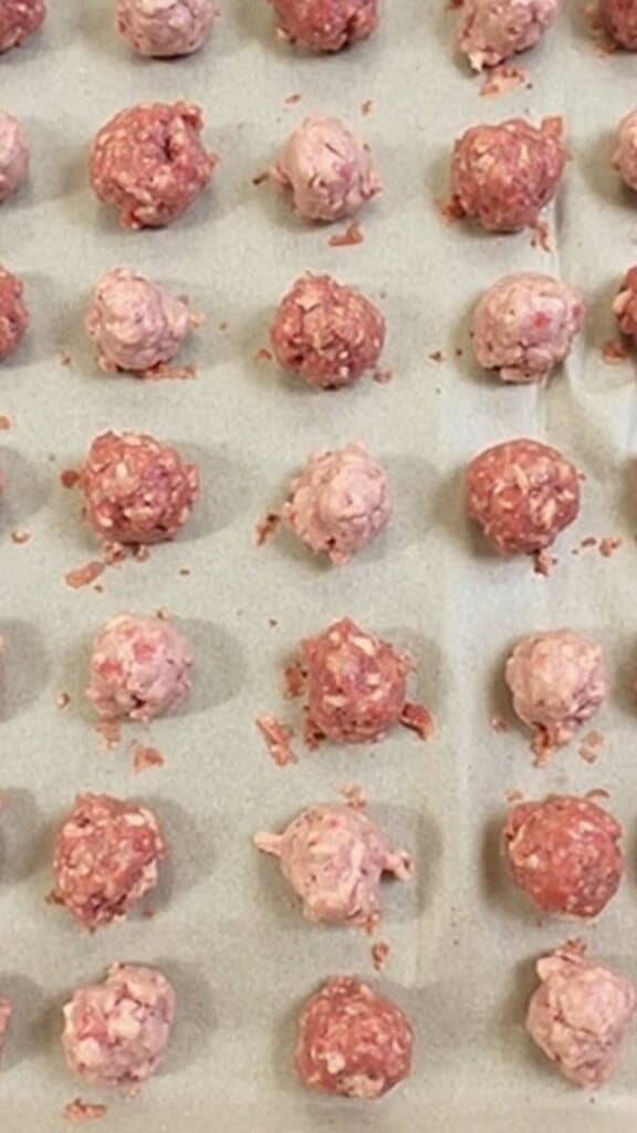 Rows of uniform pea-sized meatballs arranged in geometric patterns on a parchment lined baking sheet, ready for baking.